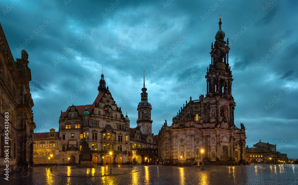 Fototapeta premium Evening view of Dresden Cathedral and historic buildings at Theater Square with illuminated baroque architecture, dramatic cloudy sky, and wet reflections in the old town of Saxony, Germany
