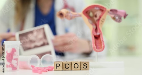 Wooden blocks on gynecology desk spell word PCOS while female doctor studies ultrasound scan. Uterus model with pills symbolize hormone disorder