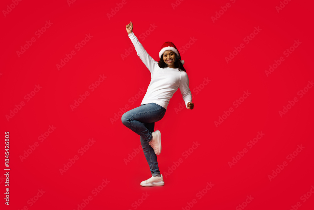 Fototapeta premium A joyful woman in casual clothing and a Christmas hat dances with excitement against a vibrant red background, celebrating the festive season with energy and happiness.