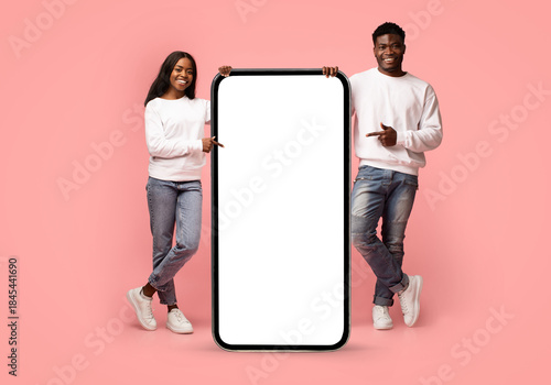 Bilde på lerret A happy millennial black couple stands in a pink studio, pointing at a big modern cellphone with an empty screen