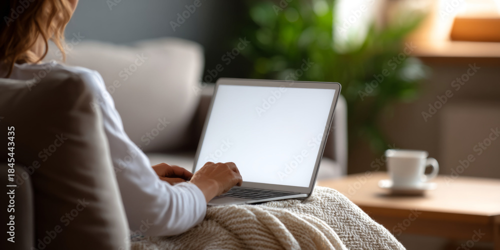 Fototapeta premium Person using laptop on cozy couch with knitted blanket in softly lit living room with coffee cup on table and green plant in background