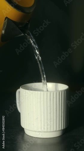 Yellow electric kettle pouring hot steaming water in modern white ceramic mug on dark countertop, perfect for preparing morning coffee or tea in kitchen. Electric kettle pouring hot water in white cup
