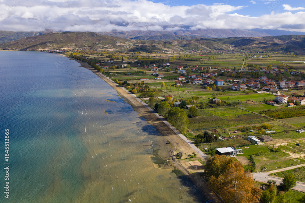 Fototapeta premium Aerial view of Pogradec lakeside with farmland, shoreline, and mountains under clouds.