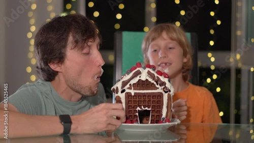Joyful moments - father and son bite into the gingerbread house they decorated with fresh berries. Cozy winter evening, family fun, creativity, and festive bonding. Slowmotion video.