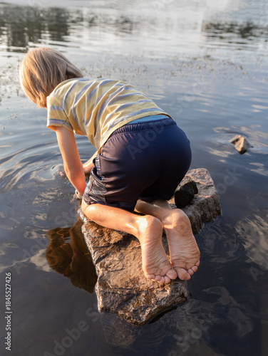 boy sits on a rock, bent over, peering into the water on the riverbank. Bare feet. Enthusiastic outdoor games as a child, weekend vacations. lifestyle
