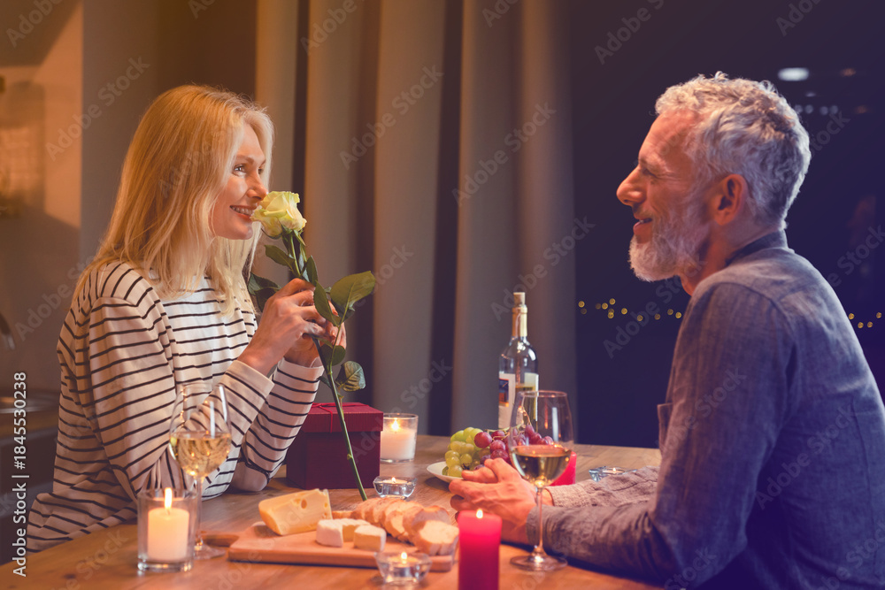 Obraz premium Happy middle-aged woman enjoying the rose fragrance during romantic dinner in the kitchen with her husband. Spouses wife and husband celebrating anniversary, special event, birthday, Valentine`s day