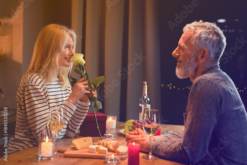 Happy middle-aged woman enjoying the rose fragrance during romantic dinner in the kitchen with her husband. Spouses wife and husband celebrating anniversary, special event, birthday, Valentine`s day