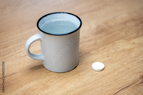 Effervescent vitamin tablet next to a mug of water on a wooden table. Fast-dissolving supplement for quick absorption, hydration support, digestion aid, and daily vitamin intake.