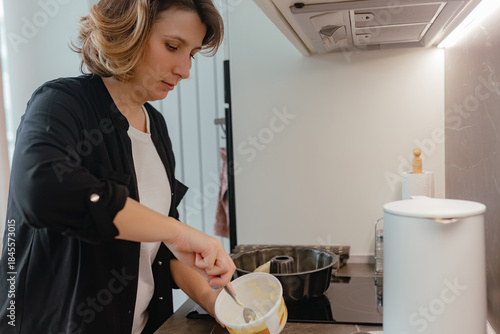 Photos Adult woman scoops margarine from yellow container while preparing mixture near stovetop area inside modern bright kitchen under warm illumination
