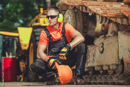 Caucasian Worker in Orange Shirt Squats Beside Heavy Machinery