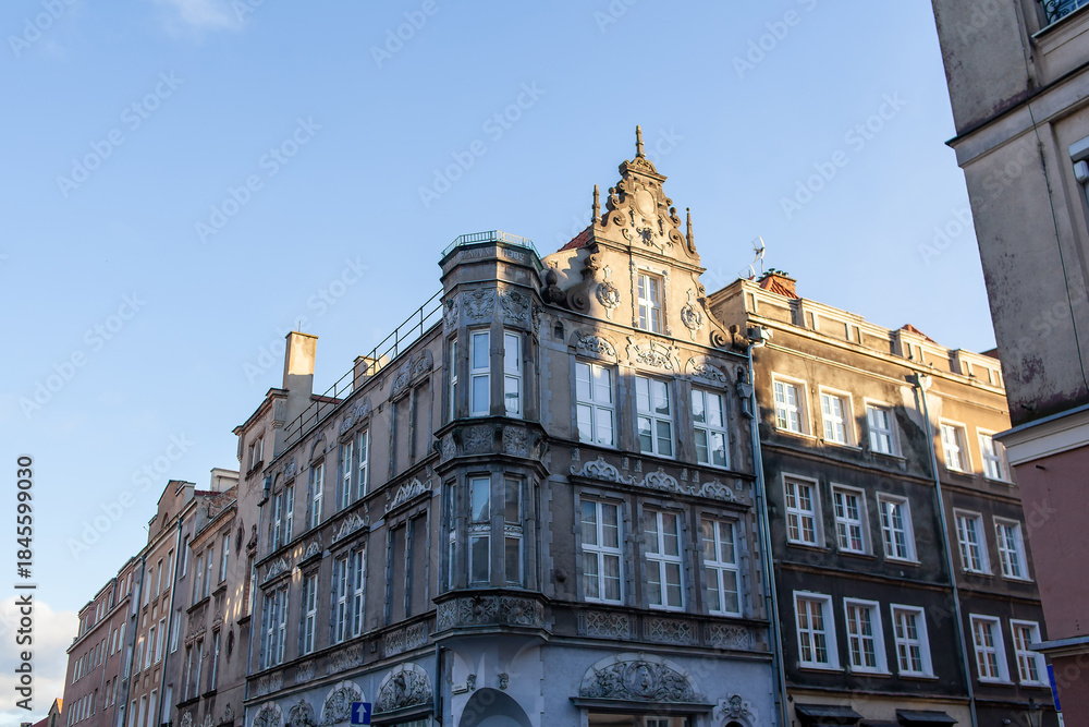 Obraz premium Old building stands tall on a busy city street, showing detailed architecture and blue sky above. Colorful buildings line the street in a historic district in Gdansk, Poland