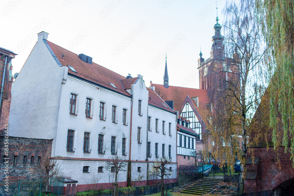 Obraz premium St Catherine's Church is the oldest church in Gdansk, Poland. A tall tower stands alongside a river while old buildings sit nearby. Trees are bare as winter approaches in the city.
