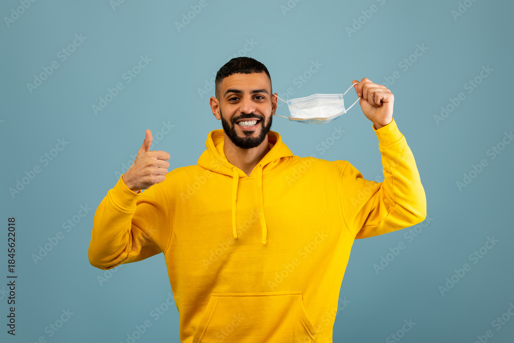 Naklejka premium A man wearing a yellow hoodie smiles broadly while waving a mask in one hand and giving a thumbs up with the other. The background is light blue, enhancing his cheerful demeanor.