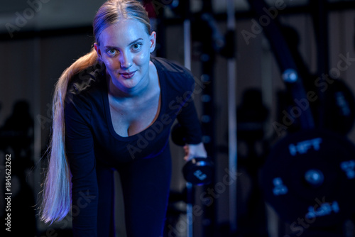 Foto Fit Caucasian woman with blonde ponytail doing dumbbell rows in dark gym