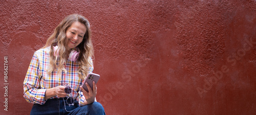 A positive mature woman with a smartphone in her hands sits against the background of a magenta color wall and listens to music.