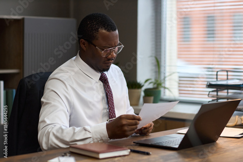 Bild auf Leinwand Middle aged Black man sitting at desk reading document, wearing glasses and form