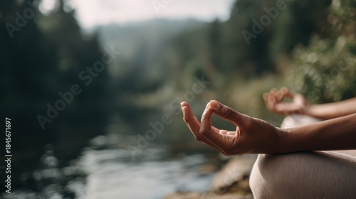 A person is meditating outdoors by a body of water with hands in a mudra gesture. This scene takes place in a natural area with soft light, creating a peaceful setting