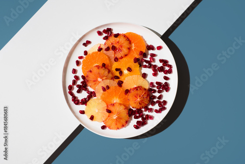 Rodajas variadas de cítricos: naranja, limón, mandarina, naranja sanguina, lima y semillas de granada, colocadas en un plato blanco sobre un fondo azul y blanco. Vista superior