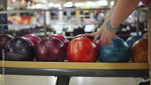 Person spins single ball on rack among many balls in bowling alley. Unidentified woman inserts fingers into the holes of orange ball and picks it up.