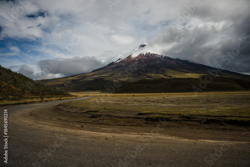 View of a majestic mountain peak capped with snow rises dramatically above a vast, open field under a sky heavy with brooding clouds, Latacunga, Cotopaxi, Ecuador.