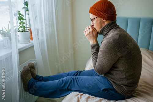 Man in beanie and sweater warming hands and feet on a radiator at home during winter, stressed about rising energy bills and trying to reduce heating costs and consumption