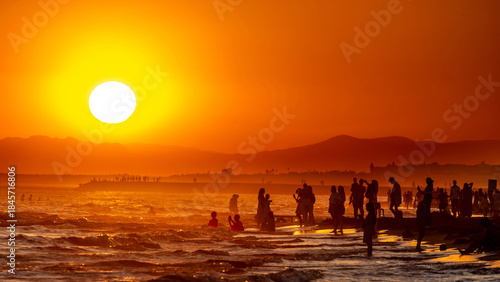 Fototapeta Naklejka Na Ścianę i Meble -  Countless silhouettes of people on a beach against a glowing orange sunset over the shimmering sea. Warm summer vacation atmosphere in backlight. Evrenseki, Turkey, Mediterranean.

