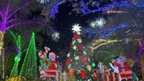 Inflatable Santa characters and a decorated Christmas tree glow with colorful lights and bows at night. Snowflake lights and drifting bubbles create a magical holiday display outdoors.