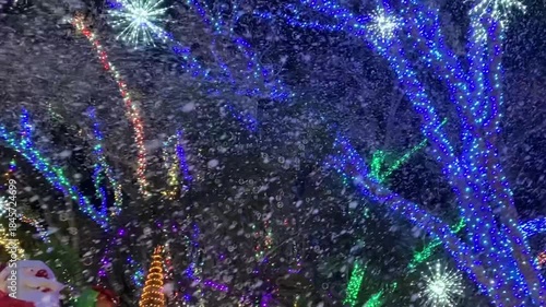 An inflatable Santa waves while holding a present near a teddy bear decoration under glowing trees. Colorful lights and floating bubbles create a festive winter scene outdoors at night.