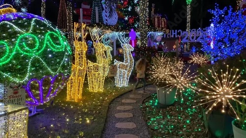 A child walks on a garden path toward glowing ornaments, reindeer lights, and sparkling decorations at night. The colorful holiday display creates a magical outdoor Christmas scene for families.