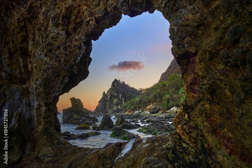 View of the rugged coast through a natural rock window, where the golden sunset kisses the horizon in Watu Lumbung Beach, Yogyakarta, Special Region of Yogyakarta, Indonesia.