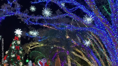 A large Christmas tree with bright lights and holiday decorations glows in a festive outdoor scene. Santa figures, ornaments, and snowflake lights fill the tropical garden with winter cheer.