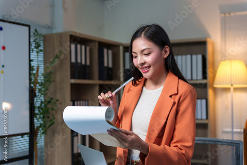 Young professional Asian businesswoman working late, reviewing documents, smiling with happiness and satisfaction in a modern office setup