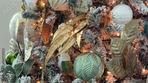 Close up of a flocked Christmas tree decorated with green and white baubles, ribbon, and warm fairy lights. Shiny ornaments create a festive holiday atmosphere indoors.