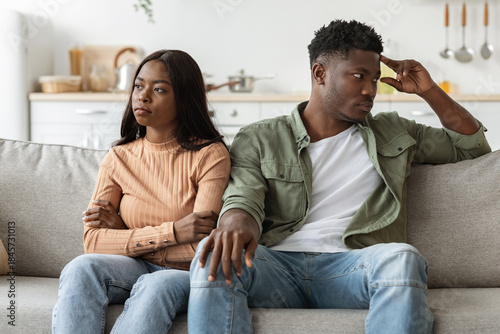 An upset African American couple sits on a couch in their living room. They are quietly processing their feelings after a quarrel, contemplating the future of their relationship.