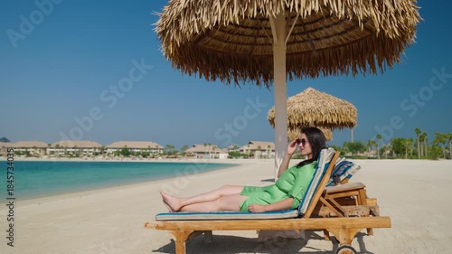 A woman in a green dress lounges on a wooden beach chair under a thatched umbrella by the turquoise sea

