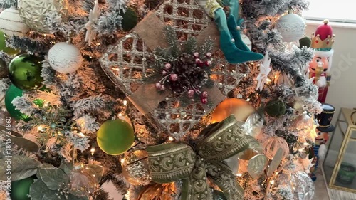 Close up of a decorated Christmas tree with a green ribbon bow, star ornaments, and mixed baubles in gold and white tones. Warm fairy lights create a cozy festive atmosphere at home.