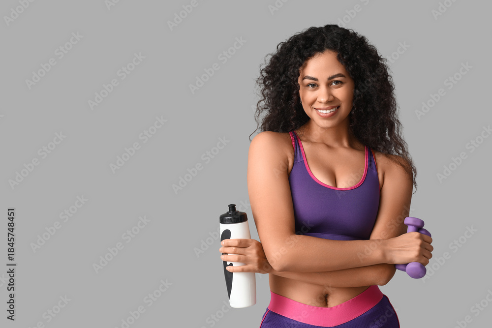 Naklejka premium Sporty young African-American female coach with water bottle and dumbbells on white background