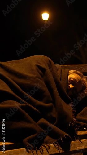 Homeless man sleeping on bench at night under street lamp wrapped in blanket peaceful sleeping person on park bench under lamp shadowed face