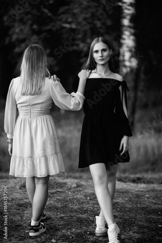Two young women standing together in a forest, dressed in contrasting light and dark outfits, showcasing friendship and connection in a serene natural environment. Black and white photo.