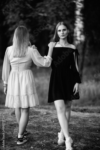Two young women standing together in a forest, dressed in contrasting light and dark outfits, showcasing friendship and connection in a serene natural environment. Black and white photo.