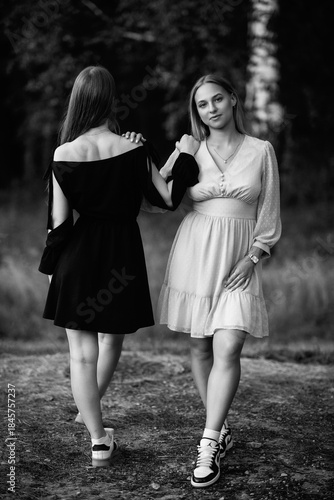Two young women standing together in a forest, dressed in contrasting light and dark outfits, showcasing friendship and connection in a serene natural environment. Black and white photo.
