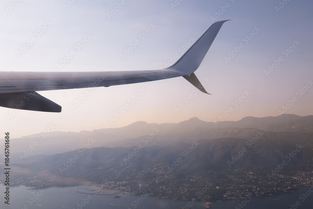 Fototapeta premium View of airplane wing over mountains and city landscape during sunset from high altitude