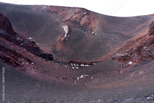 Volcanic crater of Mount Etna in Sicily, Italy. Dark lava ash slopes with red volcanic rock formations and traces of snow, showing the raw geological landscape of an active volcano.