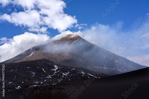 Wide view of Mount Etna volcano in Sicily, Italy, with its smoking summit partially covered by clouds. Dark volcanic slopes with patches of snow, visible hikers in the distance, and dramatic sky