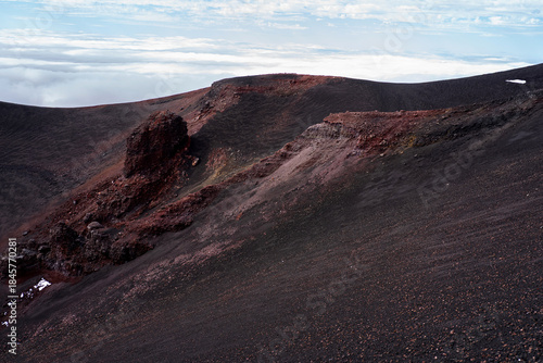 Volcanic crater ridge on Mount Etna in Sicily, Italy. Dark lava ash slopes with red volcanic rock formations rising above a layer of clouds, highlighting the dramatic landscape of an active volcano.