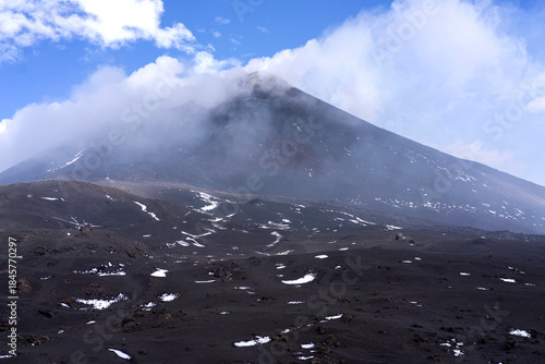 Wide view of Mount Etna volcano in Sicily, Italy, with its smoking summit partially covered by clouds. Dark volcanic slopes with patches of snow, visible hikers in the distance, and dramatic sky