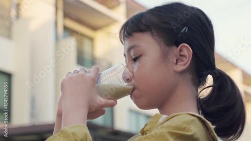 Side view of adorable Asian girl drinking fresh milk from glass outdoors. Healthy child consuming daily calcium nutrition for growth and strong bones against blurred urban building background.