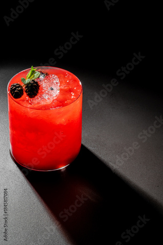 Fotografie A vibrant, red blackberry cocktail with ice cubes and fresh mint garnish in a rocks glass, dramatically lit on a dark background