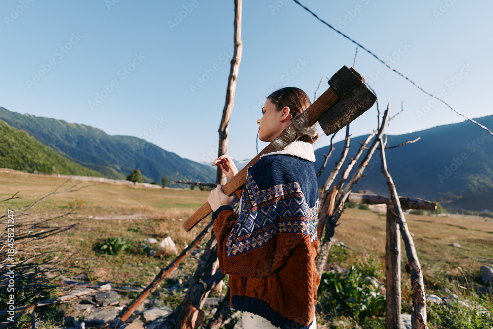 Fototapeta premium Woman with axe by fence in mountains field, wearing patterned sweater in rural wood landscape, carrying firewood over shoulder for outdoor countryside portrait and rustic lifestyle.