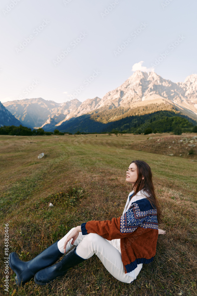 Fototapeta premium Woman sitting in a meadow wearing a patterned sweater and rubber boots, enjoying mountains and open field at golden hour, serene outdoor portrait and travel moment in nature
